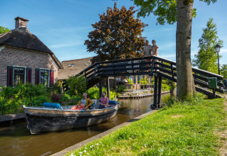 Menschen fahren mit dem Boot durch einen Kanal neben einem Ferienhaus und Brücke im Glampingpark.