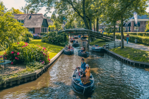 Barcas navegan por un canal en un parque de vacaciones con glamping, jardines y pintorescas casas con techo de paja.