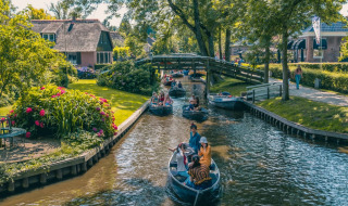 Både sejler langs en kanal ved et feriepark med glamping, omgivet af blomster, grønne træer og hytter.
