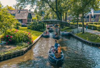 Boote fahren durch einen Kanal im Ferienpark mit Glamping-Unterkünften, umgeben von Bäumen und Blumen.