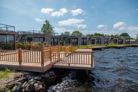 Parque vacacional con alojamientos glamping y terraza de madera junto al agua, casas modernas y cielo azul.