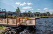 Feriepark med glampingboliger og træterrasse ved søen, moderne bygninger under blå himmel.