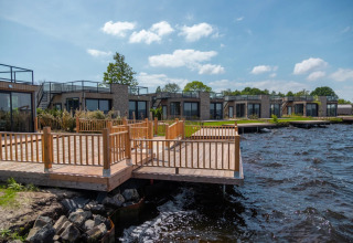 Parque vacacional con alojamientos glamping y terraza de madera junto al agua, casas modernas y cielo azul.