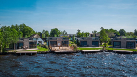 Modern glamping lodges by the lake in a holiday park surrounded by greenery and a blue sky.