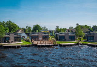 Moderne glampingboliger ved søbred i feriepark med træer og grønt landskab under blå himmel.
