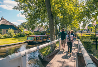 People walk on a wooden bridge beside a canal with glamping cottages and a boat in a sunny holiday park.
