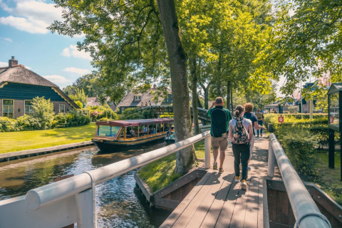 Folk går på en træbro langs en kanal med glampinghytter og båd i en feriepark på en solrig dag.