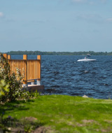 Blick auf einen See von einem Ferienpark mit Glamping, grüne Wiese, Holzzaun und Boot auf dem Wasser.