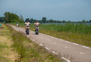 Zwei Personen fahren Elektroroller auf einer Landstraße an einem See, nahe einem Glamping-Urlaubsort.