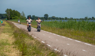Twee mensen rijden op elektrische scooters langs een meer bij een glamping vakantiepark in de natuur.