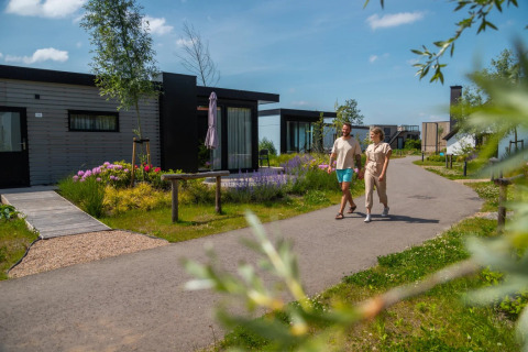 Couple walking on a path past modern glamping cabins surrounded by flowers at a sunny holiday park.