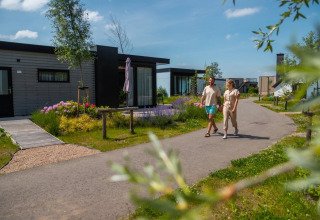 Couple walking on a path past modern glamping cabins surrounded by flowers at a sunny holiday park.
