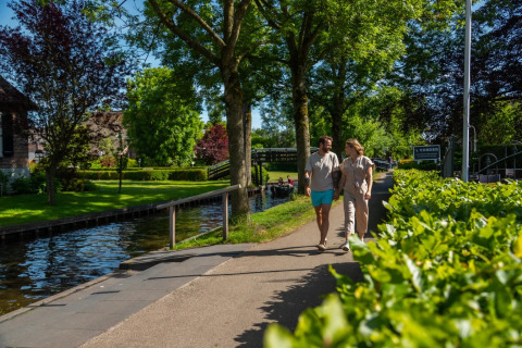 Pareja paseando de la mano junto a un canal en un parque vacacional con glamping, rodeados de vegetación.