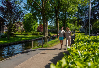 Couple marchant main dans la main près d’un canal dans un parc de vacances avec glamping, verdure luxuriante.