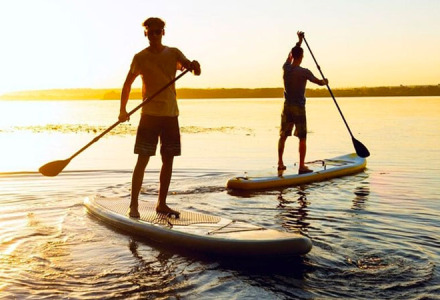 To personer står på paddleboards ved solnedgang på en sø, afslappet ferie i en glampingpark.