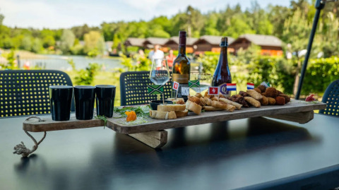 Et udendørs bord med vin, snacks og glas på et feriecenter med glampinghytter i baggrunden.