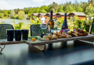 Mesa al aire libre con vino, aperitivos y vasos junto a cabañas glamping en un parque vacacional.