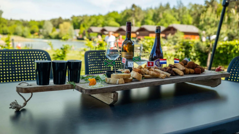 An outdoor table with wine, snacks, and glasses near lakeside glamping cabins at a holiday park.