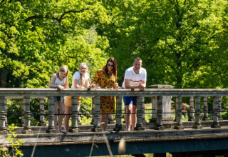 Vier Personen lehnen sich auf einer Brücke in einer grünen Ferienanlage mit Glamping-Angeboten.