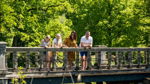 Vier Personen lehnen sich auf einer Brücke in einer grünen Ferienanlage mit Glamping-Angeboten.