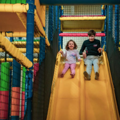 Two children laughing as they slide down a yellow slide in an indoor play area at a glamping holiday park.