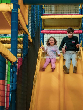 Dos niños se deslizan riendo por un tobogán amarillo en un parque de juegos interior de un glamping resort.