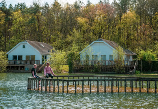 Zwei Menschen sitzen auf einem Holzsteg vor Hütten am See im bewaldeten Glamping-Ferienpark.