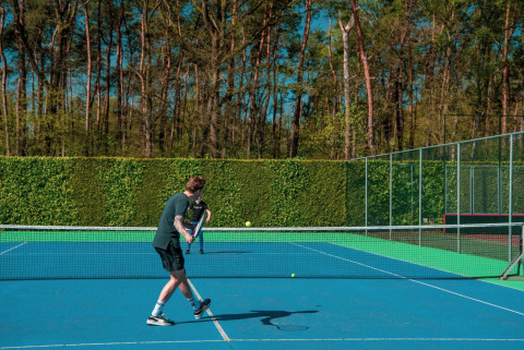 To personer spiller tennis på en udendørs bane omgivet af træer og hække under en blå himmel.