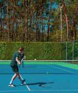 Dos personas juegan al tenis en una pista exterior rodeada de árboles y setos bajo un cielo azul claro.