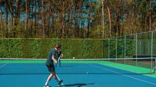 Two people playing tennis on an outdoor court surrounded by lush trees and hedges under a clear blue sky.