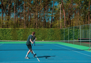 Twee mensen spelen tennis op een buitenbaan omringd door bomen en heggen onder een heldere blauwe lucht.