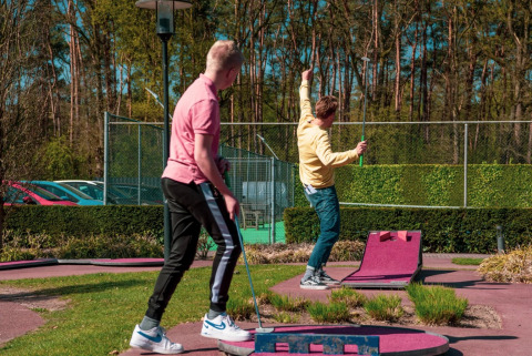 Deux jeunes hommes jouent au mini-golf au soleil dans un parc de vacances avec glamping et verdure.