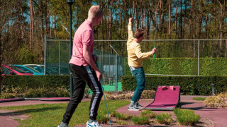 Two young men play mini-golf in the sun at a holiday park with glamping and green outdoor surroundings.