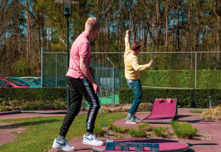 Deux jeunes hommes jouent au mini-golf au soleil dans un parc de vacances avec glamping et verdure.