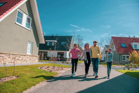 Familie spaziert auf dem Weg zwischen modernen Ferienhäusern im Ferienpark bei sonnigem Wetter.