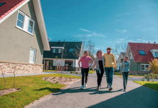 Familie gåtur på sti mellem moderne sommerhuse i feriepark på en solrig dag med blå himmel.
