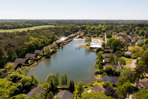 Luftaufnahme eines Ferienparks mit See und Glamping-Unterkünften, umgeben von viel Grün und Hütten.