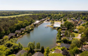 Aerial view of a holiday park with a central lake, surrounded by glamping lodges and lush green trees.