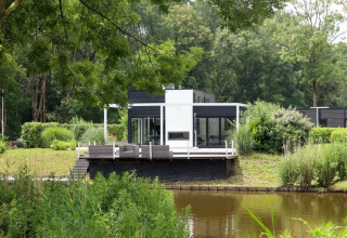 Cabane de glamping moderne au bord d’un lac dans un parc de vacances, entourée de verdure et d’arbres.