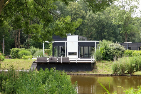 Cabane de glamping moderne au bord d’un lac dans un parc de vacances, entourée de verdure et d’arbres.