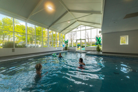Piscina cubierta con grandes ventanales y tres personas nadando en un parque vacacional con glamping.