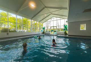 Indendørs swimmingpool med store vinduer og tre personer der svømmer på et feriecenter med glamping.