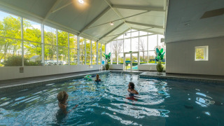 Piscina cubierta con grandes ventanales y tres personas nadando en un parque vacacional con glamping.