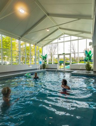 Piscina cubierta con grandes ventanales y tres personas nadando en un parque vacacional con glamping.