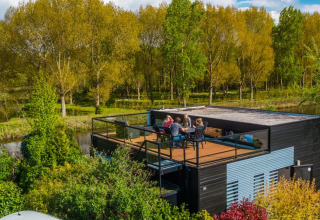 Tagterrasse med gæster ved moderne glampingbolig i feriepark, omgivet af frodige træer og sø.