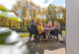 Familie sidder udenfor ved bord, nyder solskin ved en feriepark med glamping, omgivet af træer.