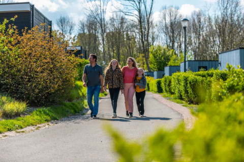 Family walking on a path at a holiday park surrounded by greenery and glamping cabins on a sunny day.