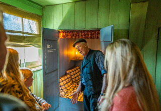 A guide shows a traditional box-bed to visitors inside a rustic holiday park with glamping cabins.