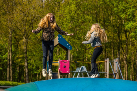 Deux filles jouent et sautent sur un grand coussin gonflable dans une aire de jeux entourée d’arbres.