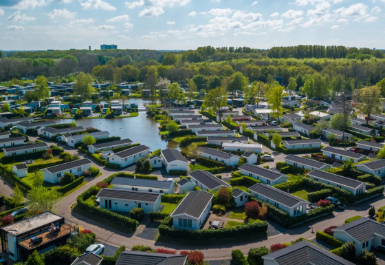 Luftfoto af et feriepark med moderne glamping-hytter, grønne områder og en sø omgivet af træer.